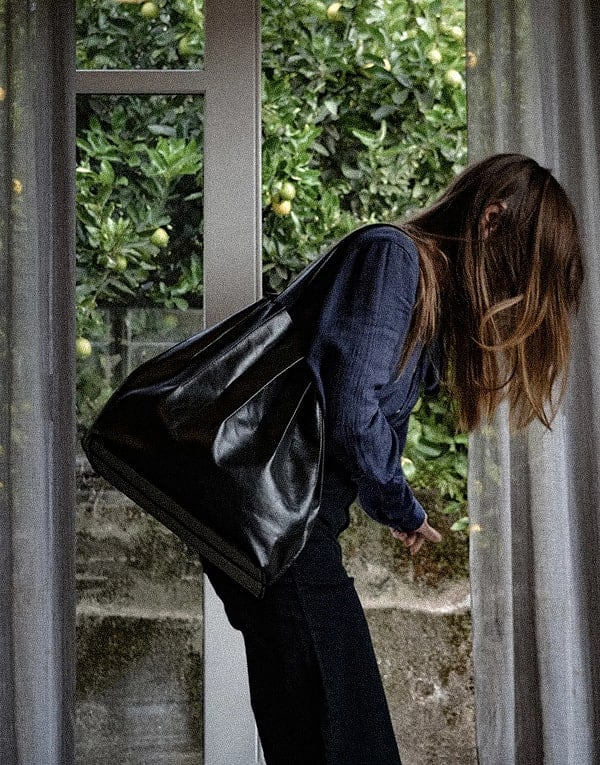 Woman with long hair leaning forward while holding a large black bag, standing by a window with visible greenery outside.