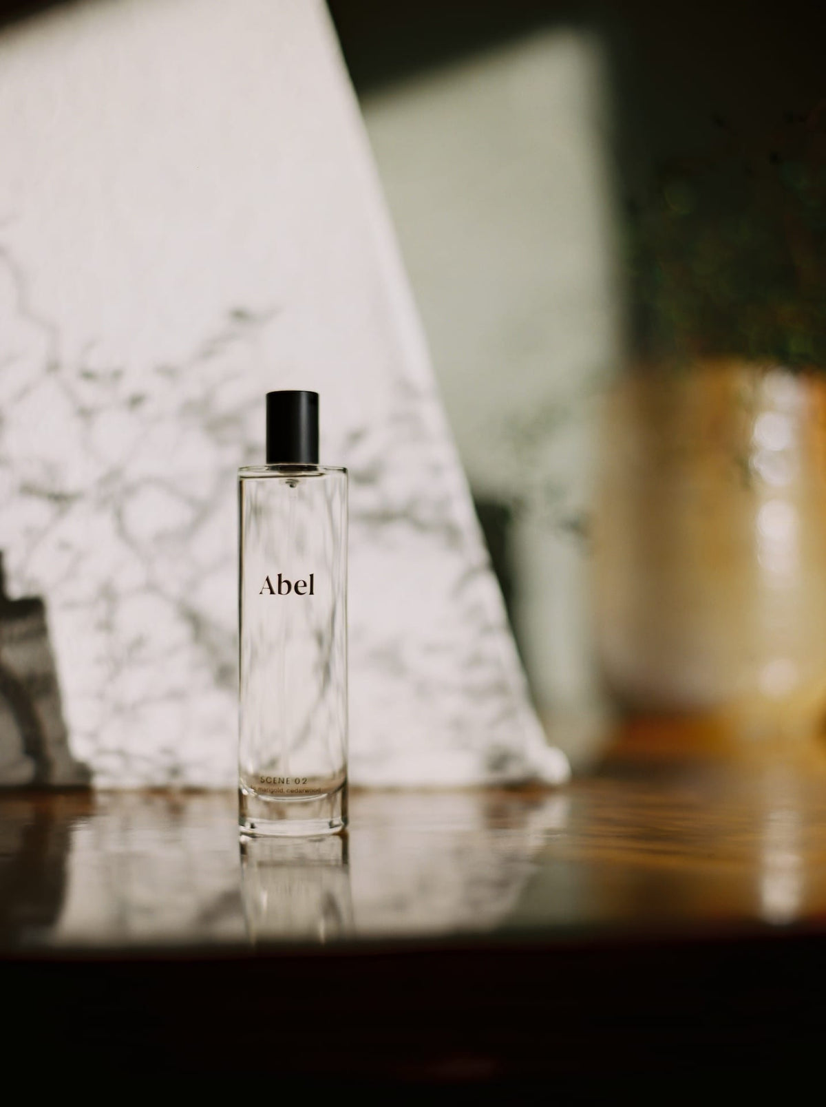 A bottle of Abel Room Spray - Scene 02 ⋅ fig, marigold, cedarwood on a reflective surface with a blurred background.