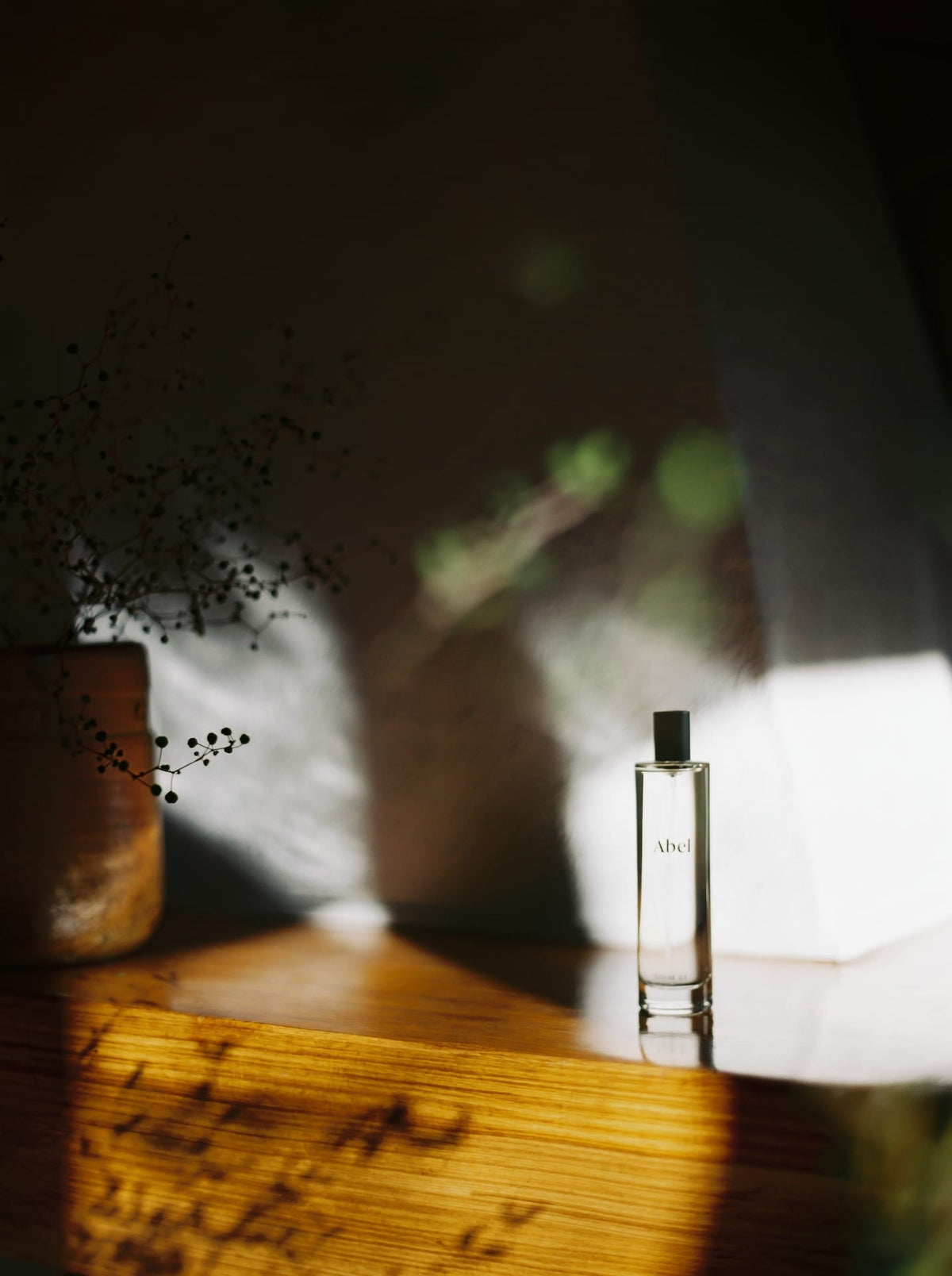 A bottle of Abel Room Spray – Scene 02 ⋅ fig, marigold, cedarwood on a wooden surface in natural lighting with shadows and a vase with dried flowers in the background.