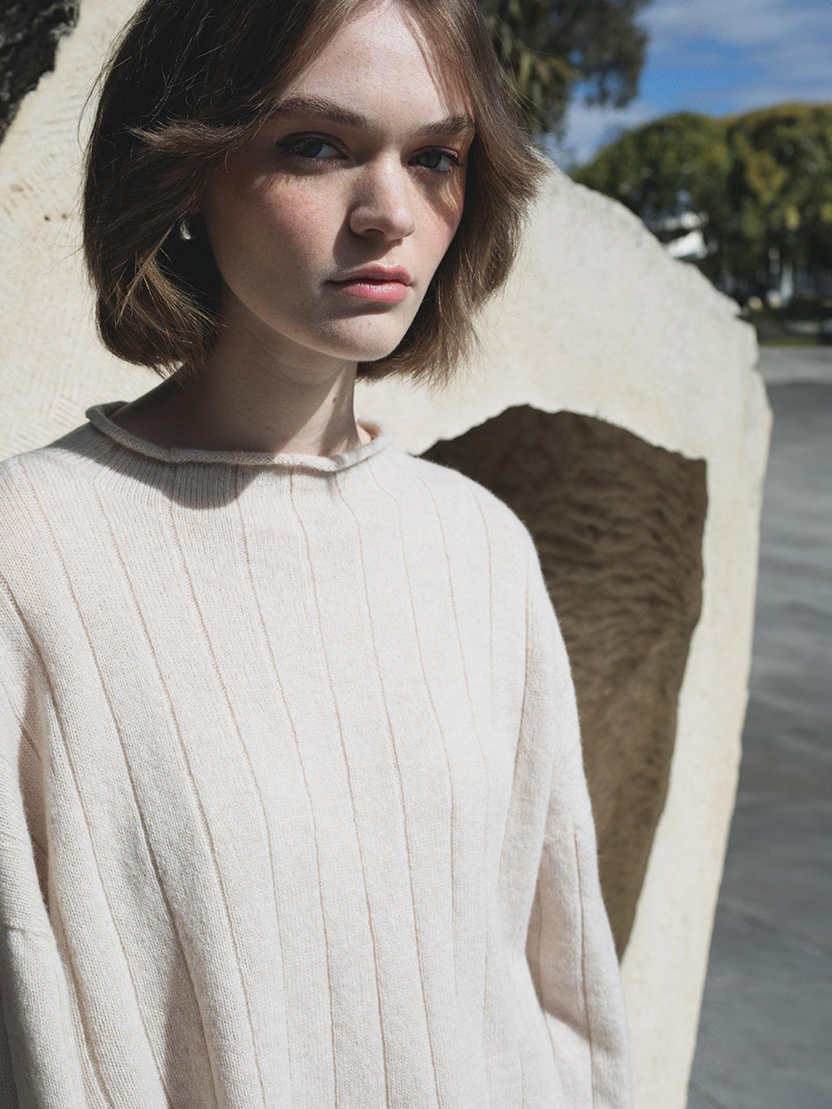 A woman in an oversized Echo Knit – Creme sweater, made from Italian air-spun merino wool, is standing in front of a rock.
