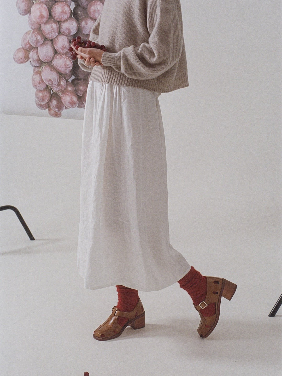Person in a beige sweater and white skirt holding grapes, with Francie Possum Merino Socks – Crimson and brown shoes (refer to size chart for details), standing in a studio setting.