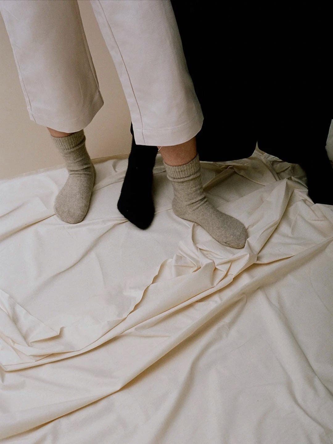 A person standing on a wrinkled beige sheet wearing one black shoe and one Francie Possum Merino Sock – Natural.