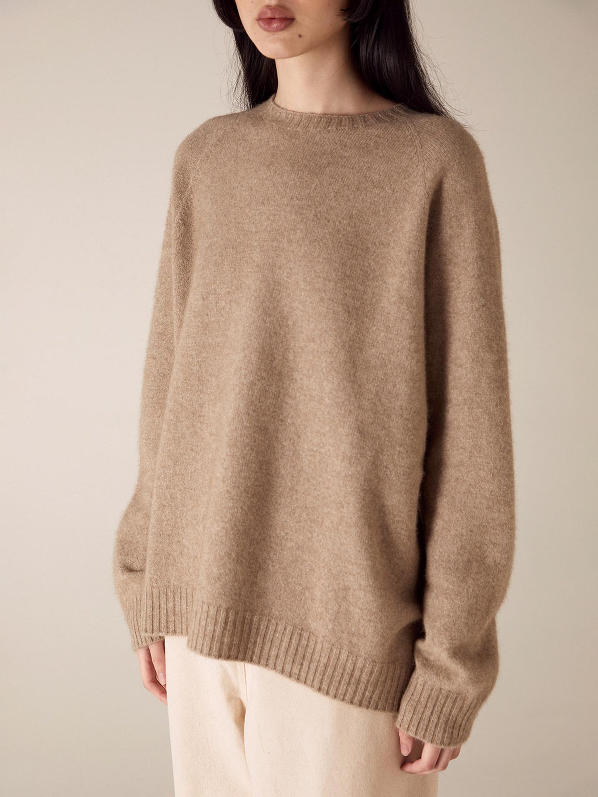 A person in a Francie Nimbus Raglan Knit - Natural sweater with a relaxed fit stands against a neutral background, focus on the texture and fit of the sweater.