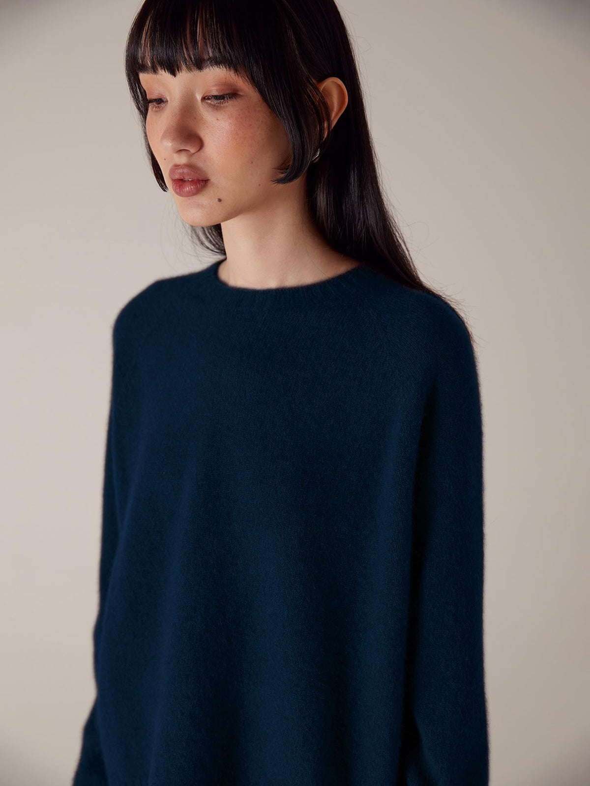 A petite woman with bobbed hair wearing a Francie Nimbus Raglan Knit in Midnight, looking downward in a softly lit setting.