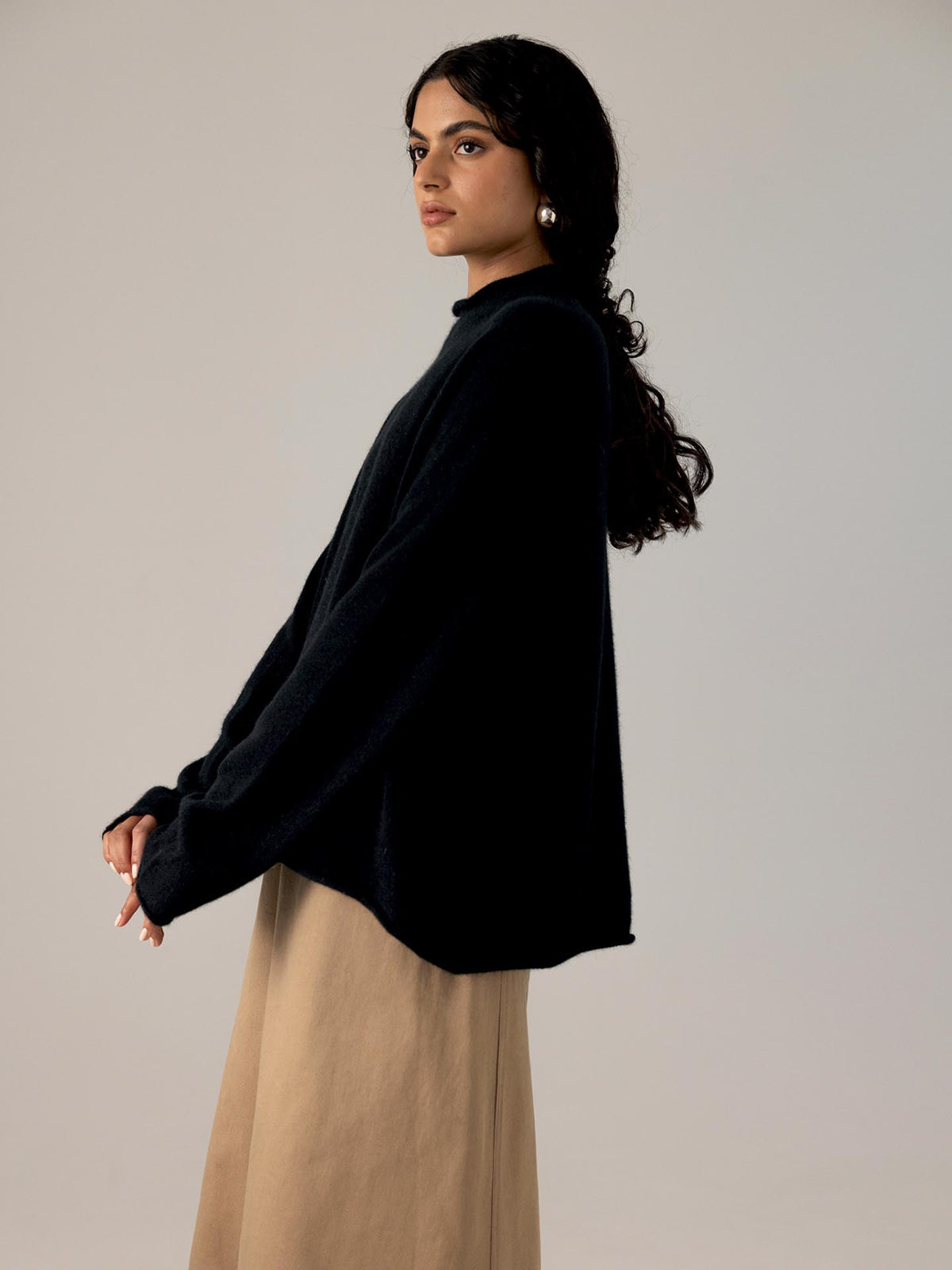 A woman in a relaxed fit Francie Eclipse Knit – Black sweater and beige skirt, standing in profile, looks over her shoulder against a neutral background.