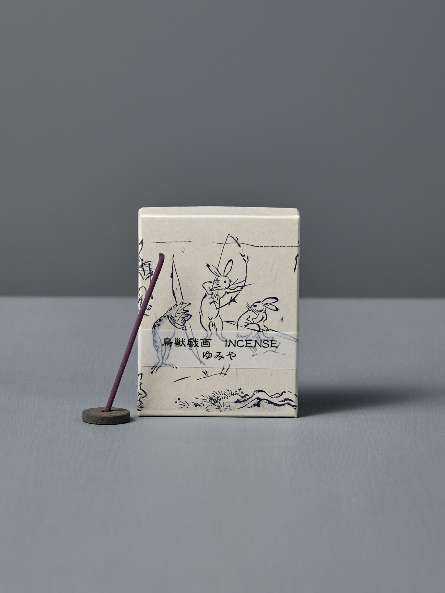 A box of Japanese Incense Yumiya – Warm Wood & Gentle Spice, featuring Japanese text and Choju Jinbutsu Giga-inspired art, stands behind a lit purple stick on a holder, releasing subtle woody fragrance against a gray backdrop.