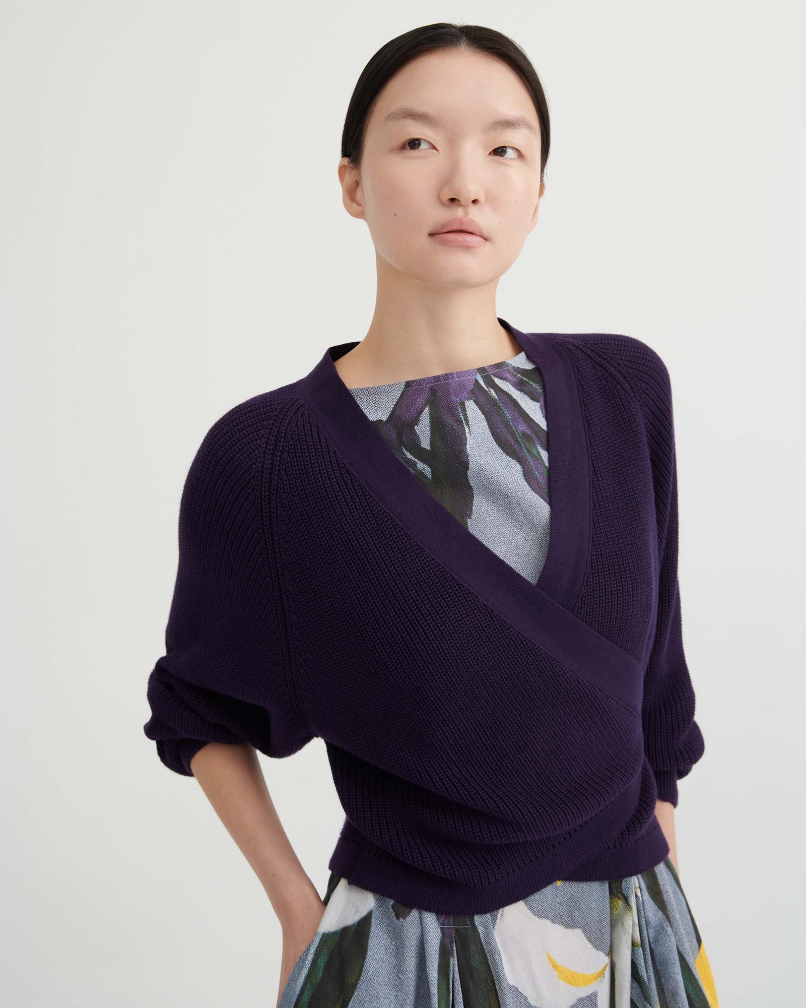 A person poses against a plain background wearing the Composure Cardigan – Dahlia by Kowtow in a relaxed fit, layered over a floral-patterned dress with a gray base and yellow accents.