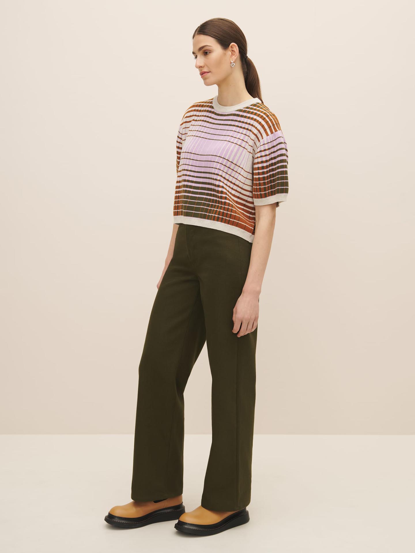 A woman stands in a neutral backdrop, wearing a striped, multicolored oversized Kowtow Gradient Knit Top with drop shoulder sleeves and dark green trousers, paired with black loafers.