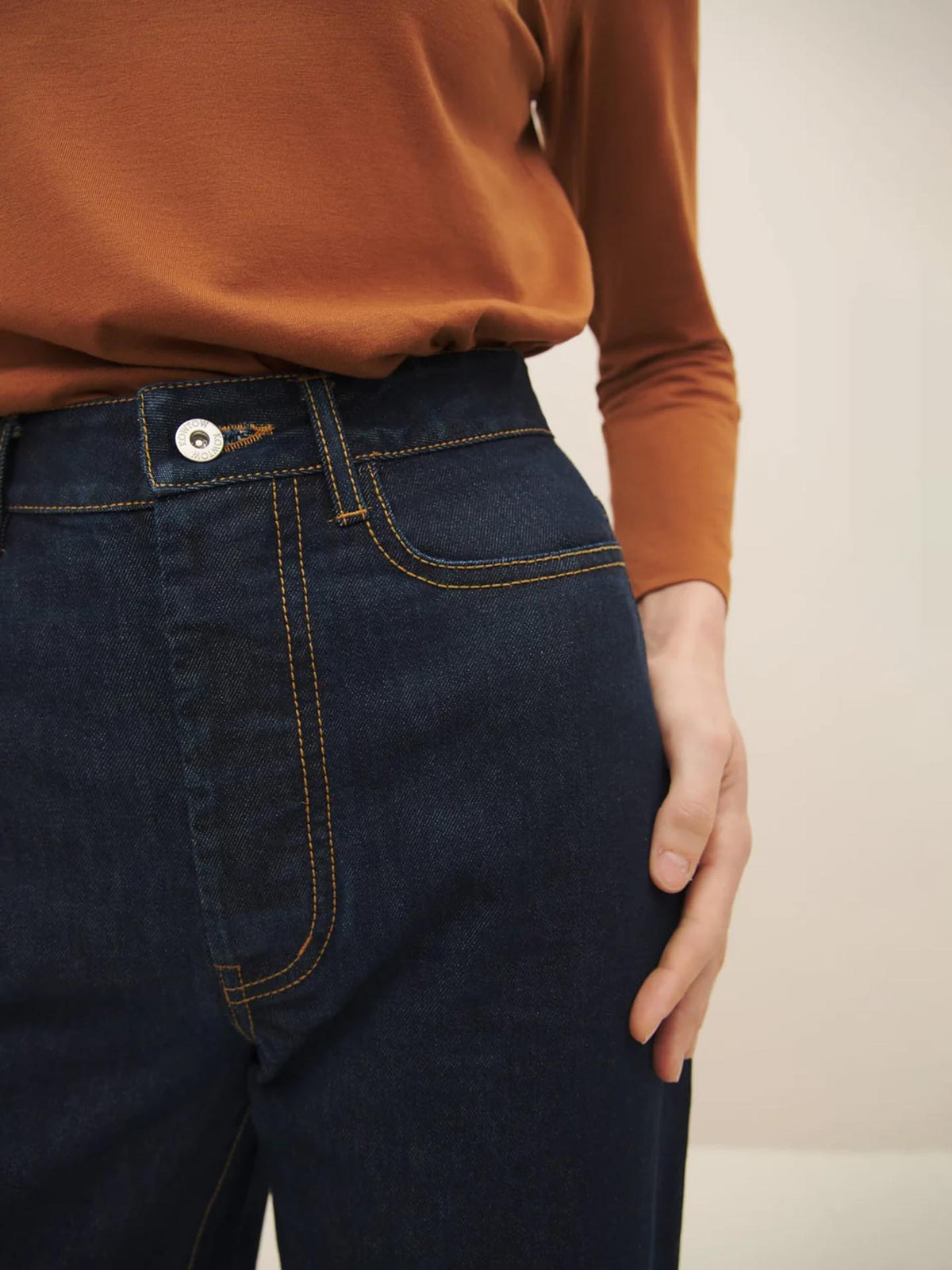 Close-up of a person wearing Kowtow high puddle jeans in indigo denim with yellow stitching, focusing on the top front part of the jeans. The person is wearing a brown top and has their hand on their hip.