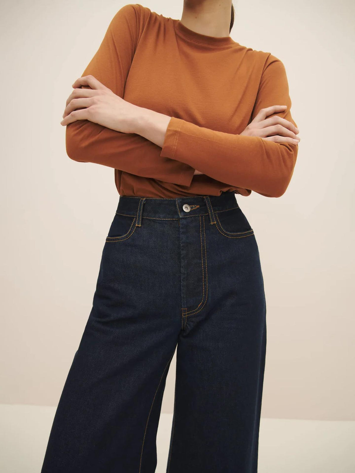 Person wearing a burnt orange long-sleeve shirt and Kowtow&#39;s High Puddle Jeans in Indigo Denim, standing with arms crossed. Only the torso and legs are visible.
