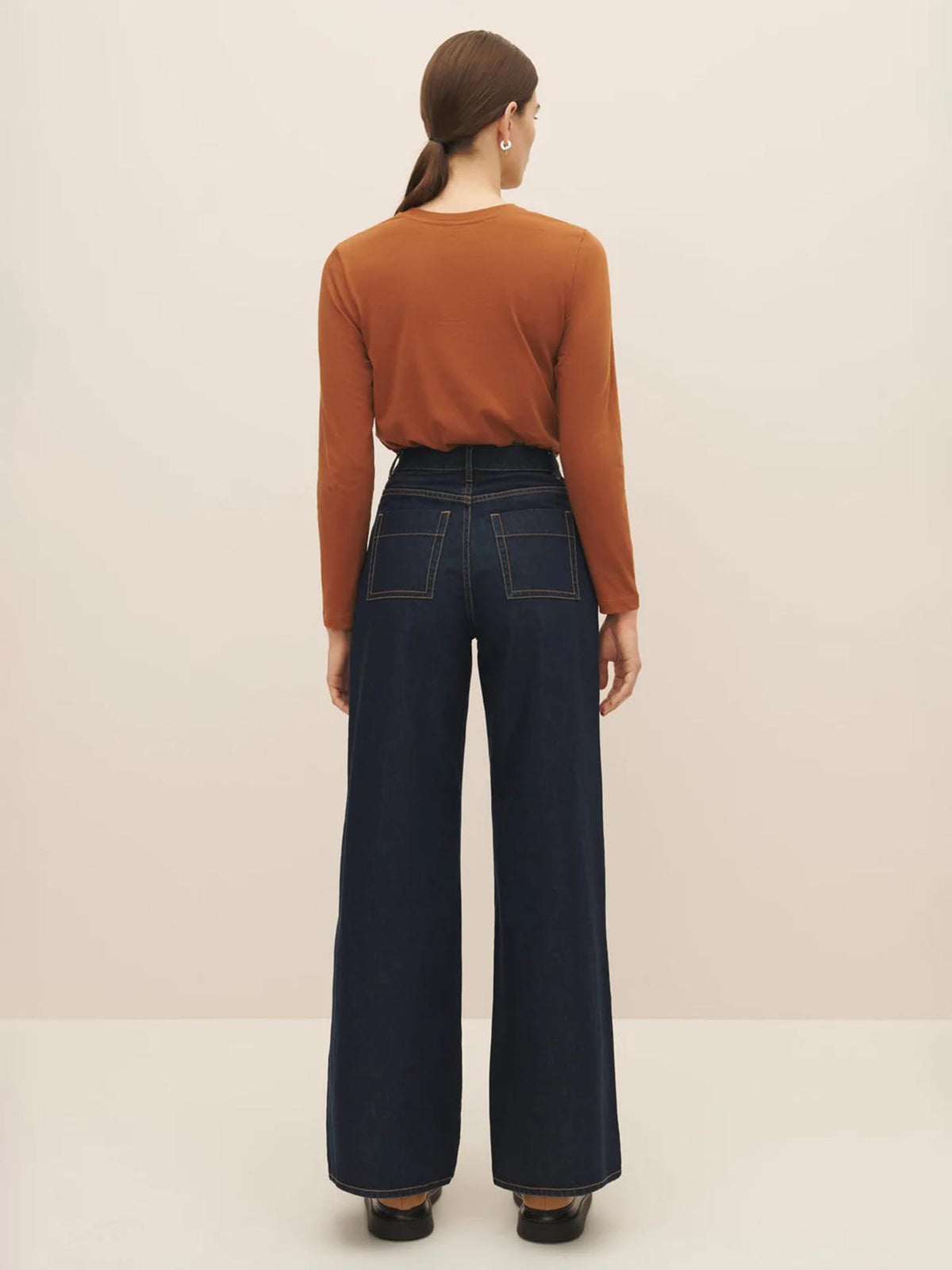 A woman viewed from the back wearing a rust-colored top and Indigo Denim High Puddle Jeans, standing against a light beige background by Kowtow.