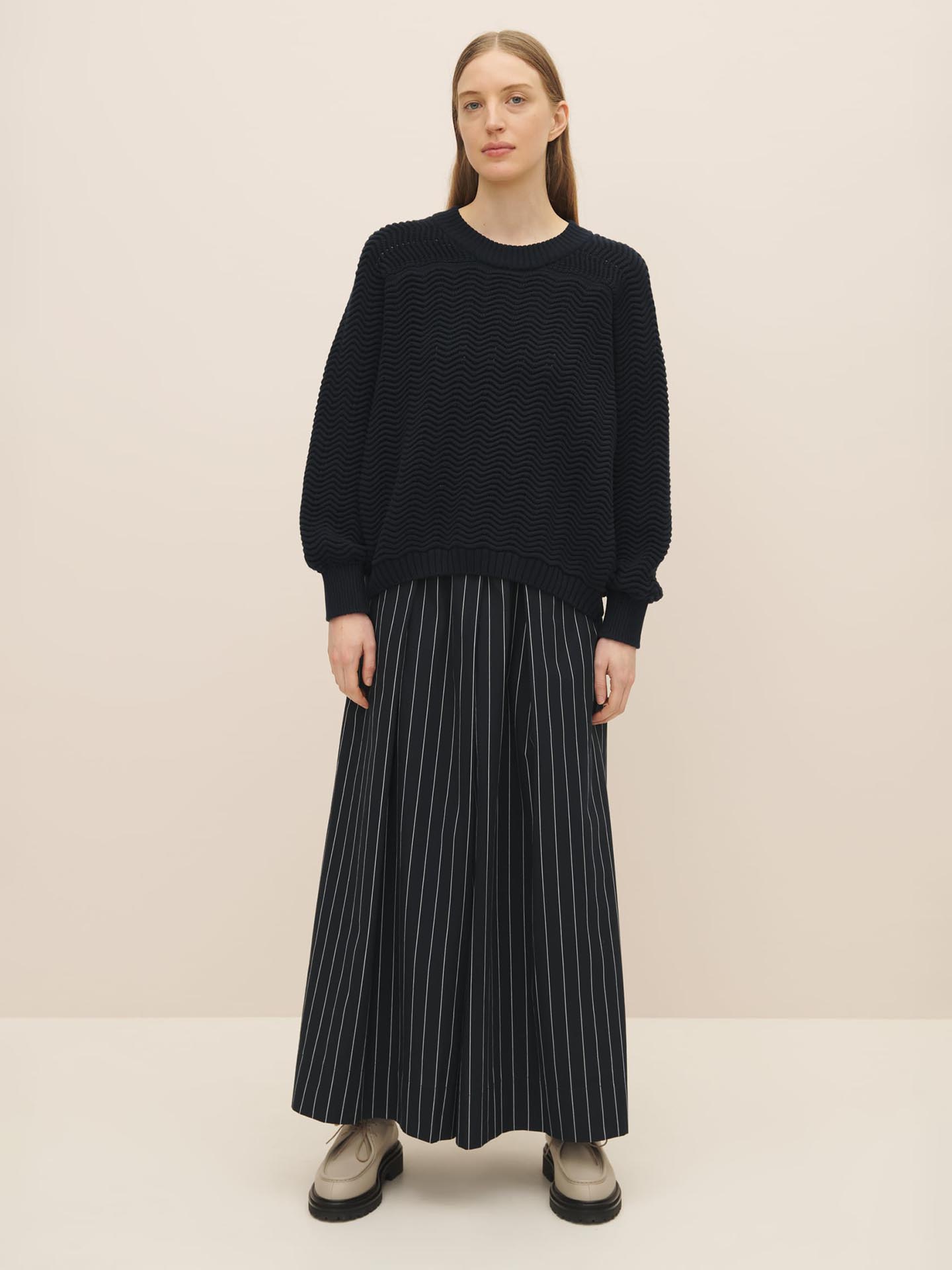 A woman in a Kowtow Zig Zag Crew – Indigo sweater and striped skirt stands against a pale background, wearing white platform shoes.
