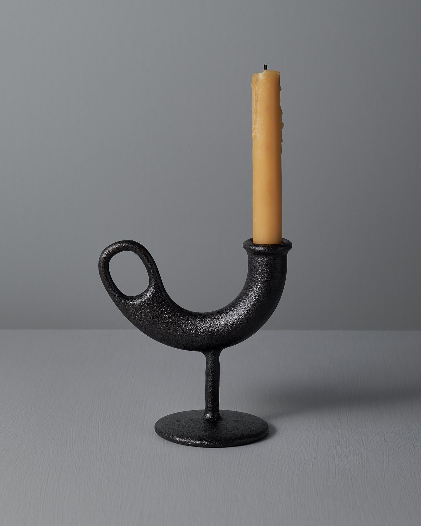 The Ildhane Candle-stick Holder by Nedre Foss is a modernist black, curved metal piece featuring a circular base, elegantly displaying a tall, unlit yellow candle against a plain gray background.