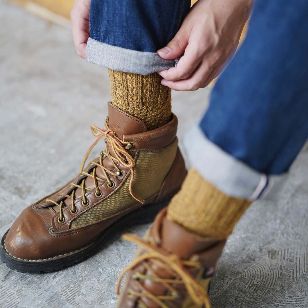 Person adjusting the cuff of jeans, revealing Nishiguchi Kutsushita Wool Cotton Boot Socks – Mustard tucked into brown leather lace-up boots. Check our size chart to find the perfect fit in both men's and women's sizes.