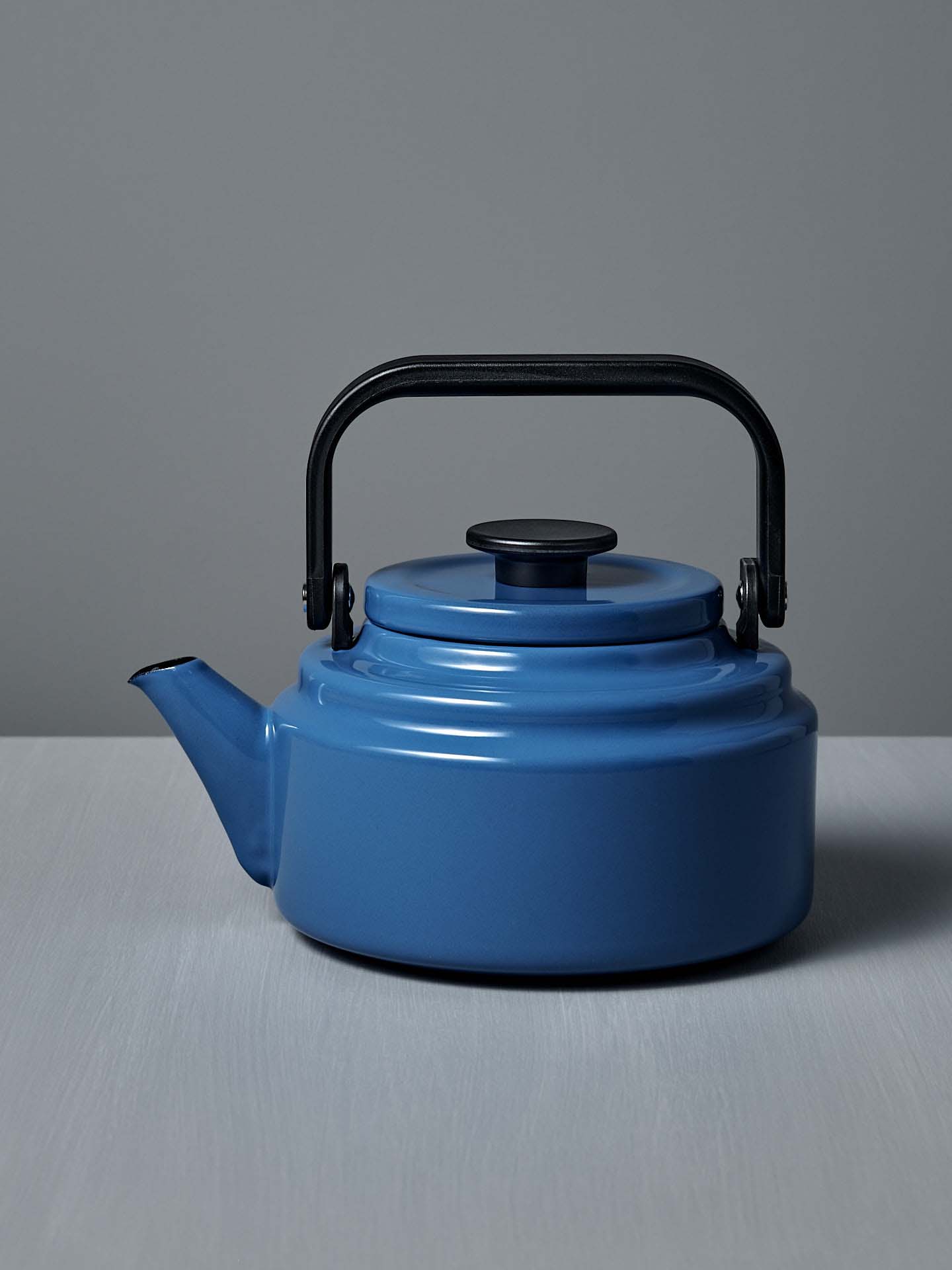 The Amu Stove-top Kettle – Blue by Noda Horo, featuring an enamelware finish and black handle and spout, sits on a gray surface against a gray background, evoking a classic Japanese design.