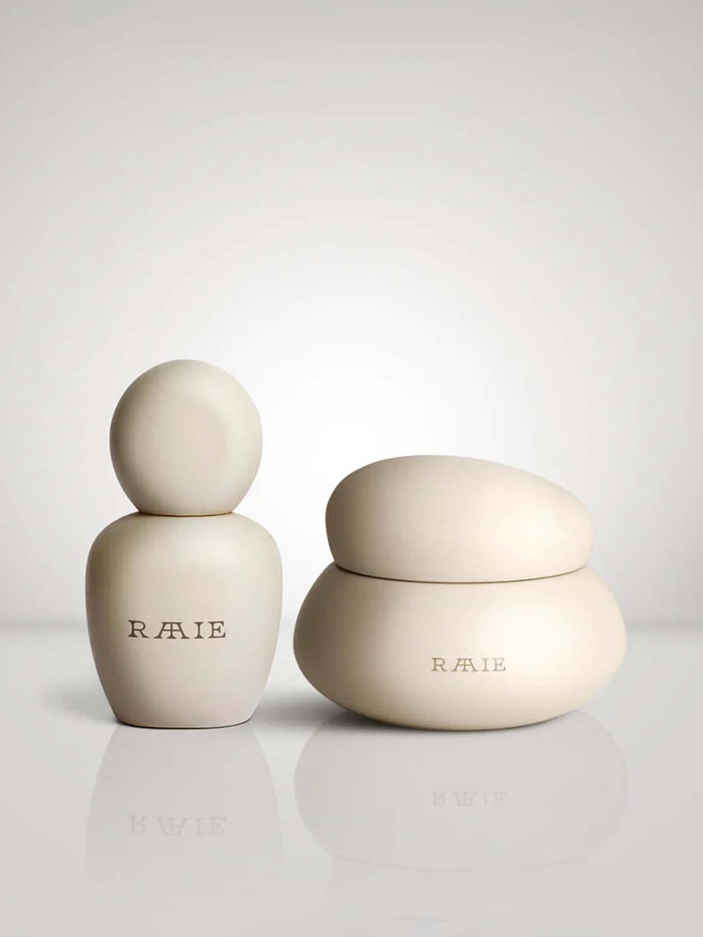 Two beige, uniquely shaped cosmetic containers with the brand name "RAAIE" boast their Bakuchiol-infused formula on a reflective surface against a plain background.