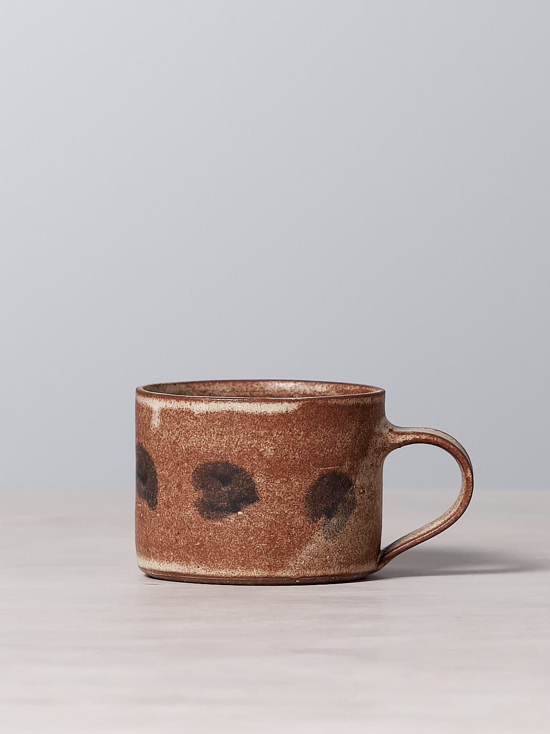 A brown Spots mug made of iron-rich clay with a matte cream glaze sits on a table.
