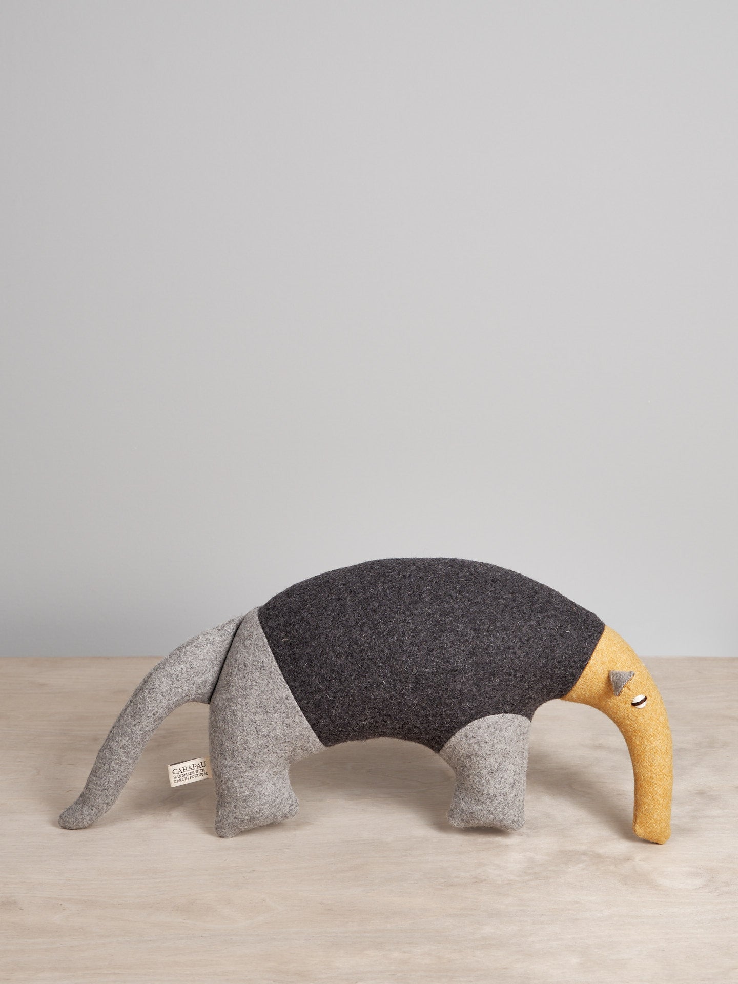 A grey and yellow Oscar, the Northern Tamandua stuffed animal on a wooden table by Carapau.