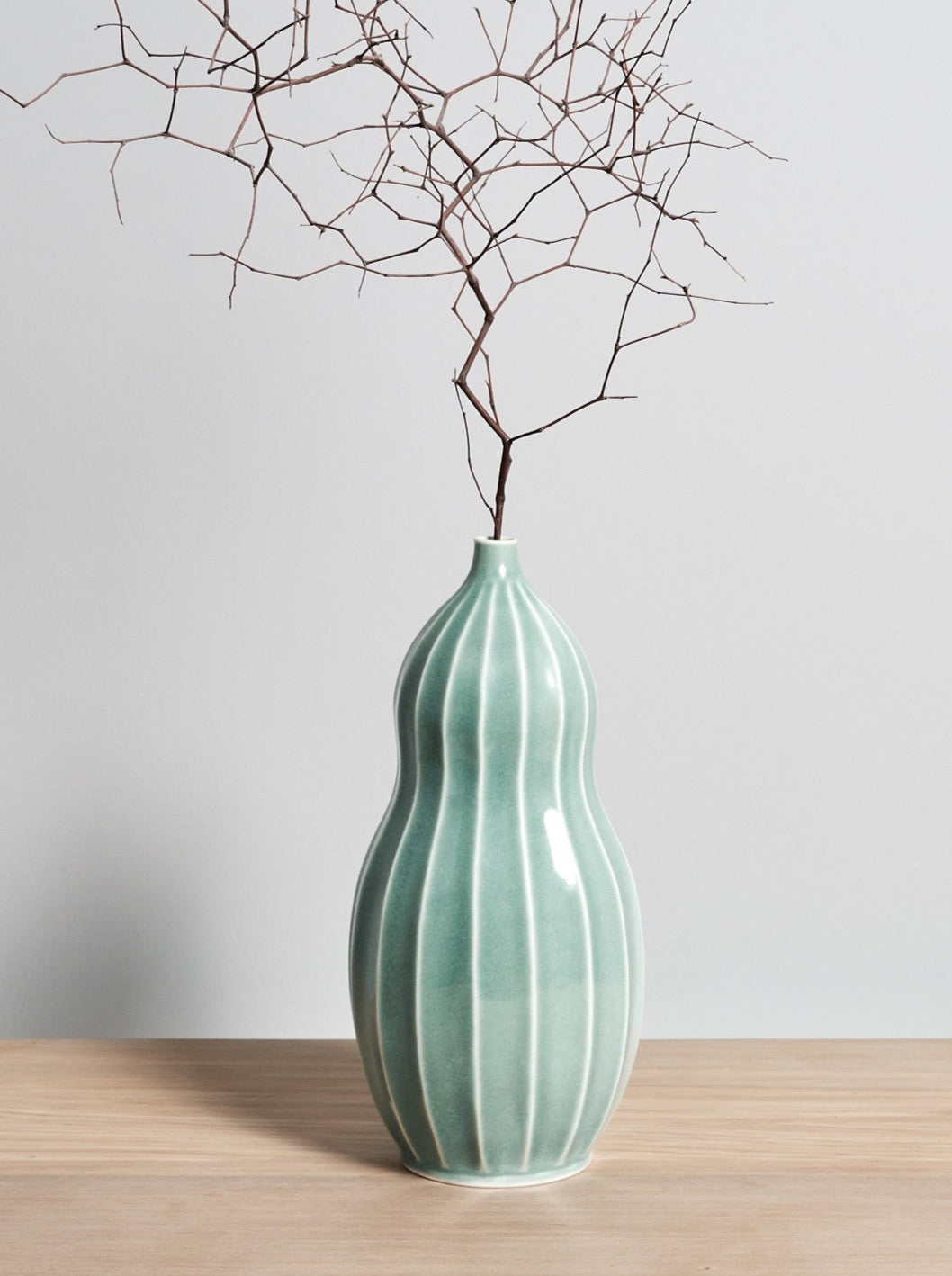 A Gourd Vase - Celadon by Jino Ceramic Studio with a twig in it.