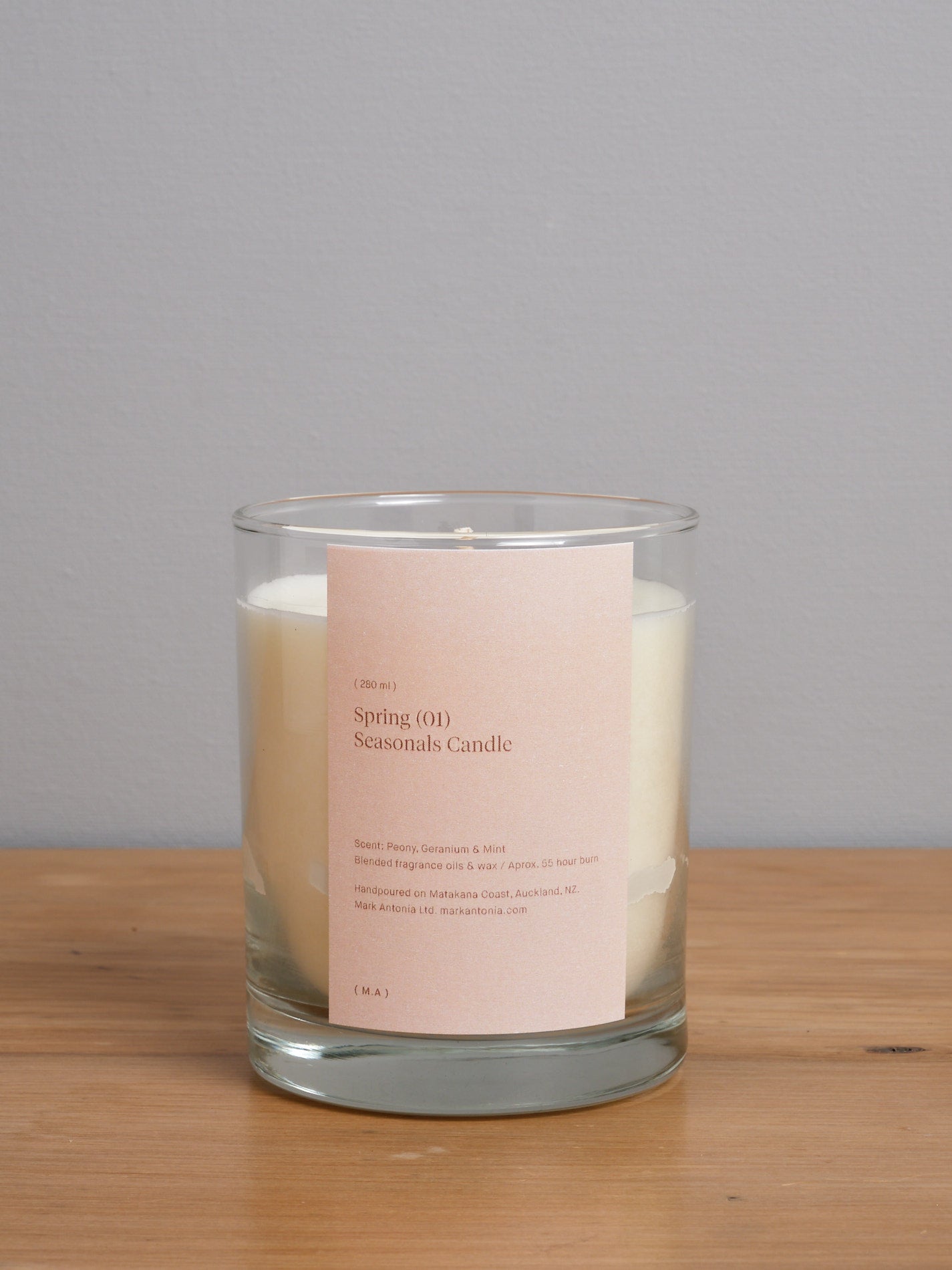 A Mark Antonia Seasonal Candle – Peony, Mint & Geranium with a label on it sitting on a wooden table.