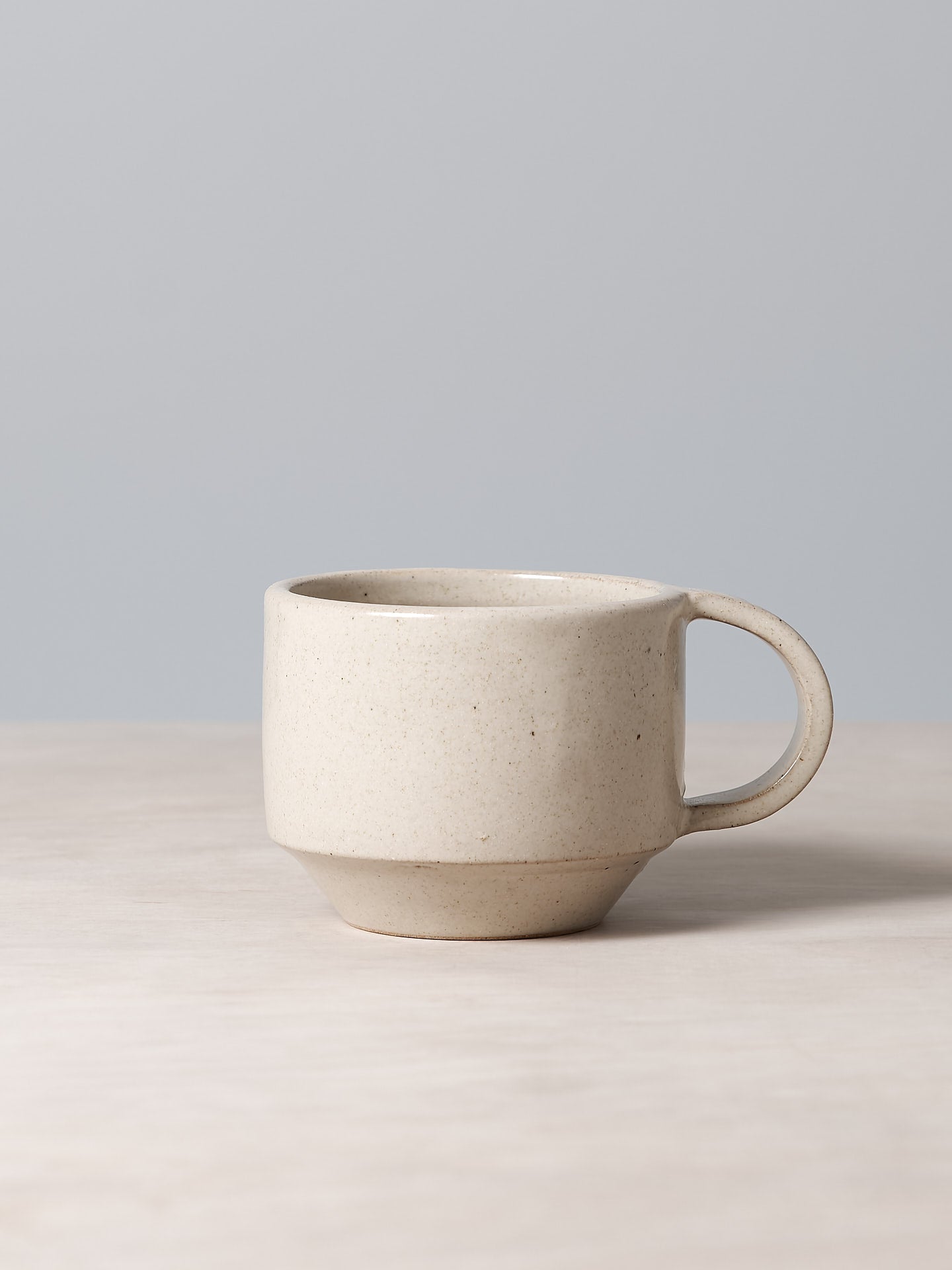 A handmade Richard Beauchamp beige coloured glaze ceramic C-handled stacking mug sitting on top of a table.