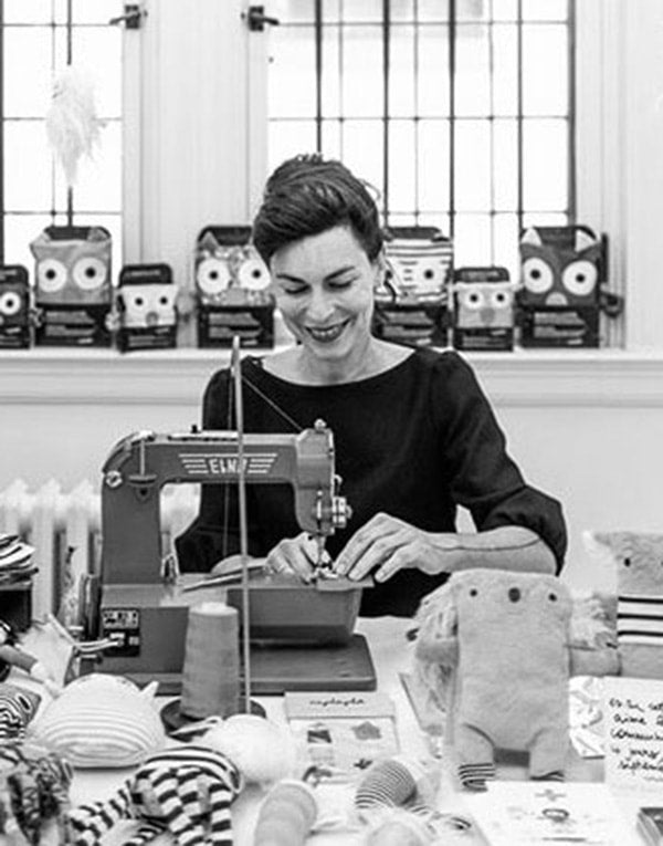 A woman sits at a sewing table with stuffed animals.