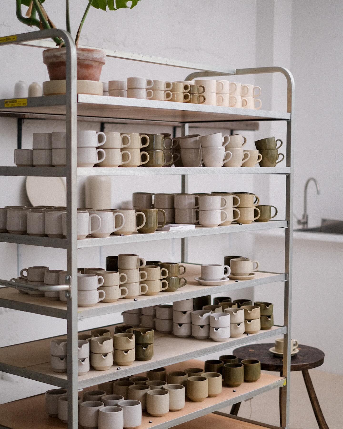 A metal rack with several shelves holds rows of neatly arranged ceramic mugs and cups in neutral colors. A potted plant sits on the top shelf. A sink is visible in the background.