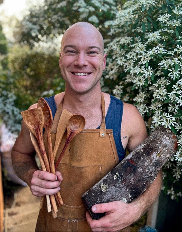 A man wearing a brown apron, holding several wooden spoons in one hand and a log in the other, stands in front of blooming white flowers.