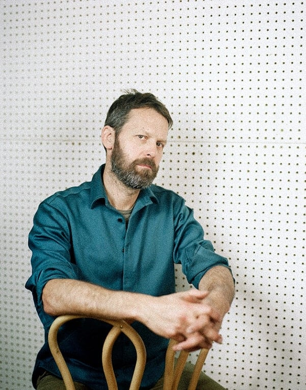 Man with a beard sitting on a chair against a pegboard background.