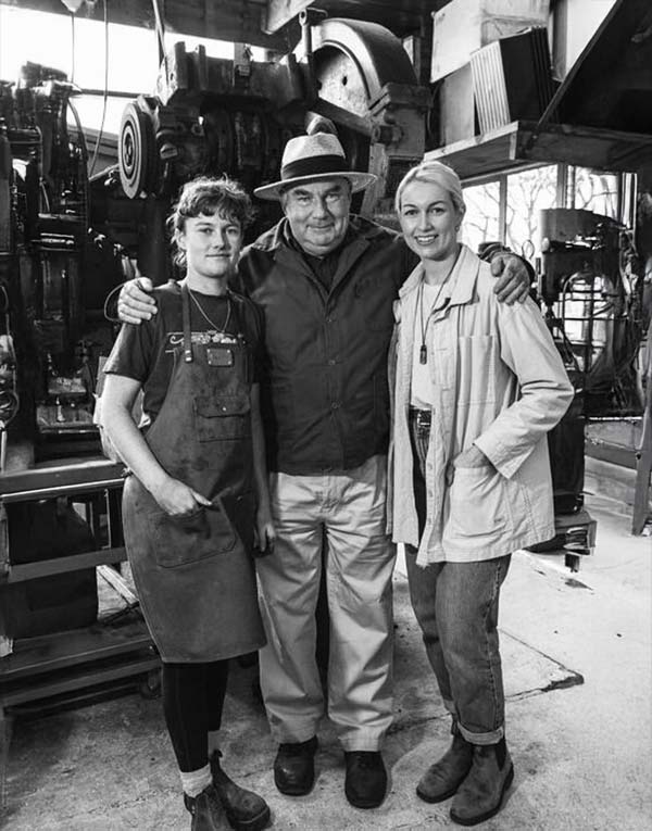 Three people stand together in a workshop, with machinery in the background. The man in the center has his arms around the two women beside him. All are looking at the camera and smiling.