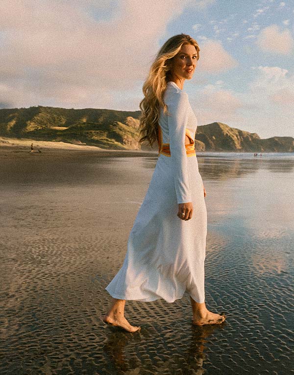 Person with long blonde hair in a white dress walking barefoot on a wet beach. Hills and ocean are in the background.