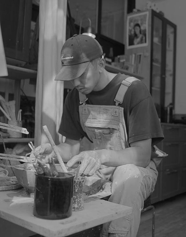 A person wearing overalls and a cap sits at a table, working with clay and brushes in an art studio.