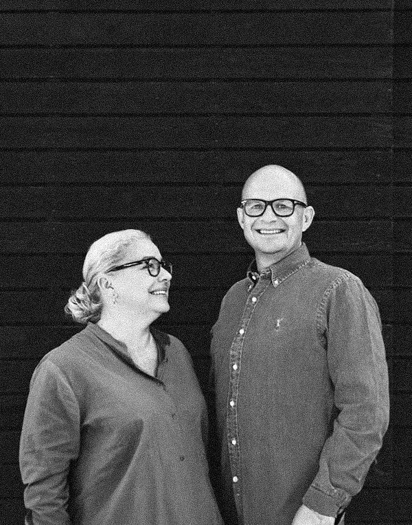 Black and white portrait of a smiling bald man and a woman wearing glasses, standing in front of a dark, horizontal striped background.