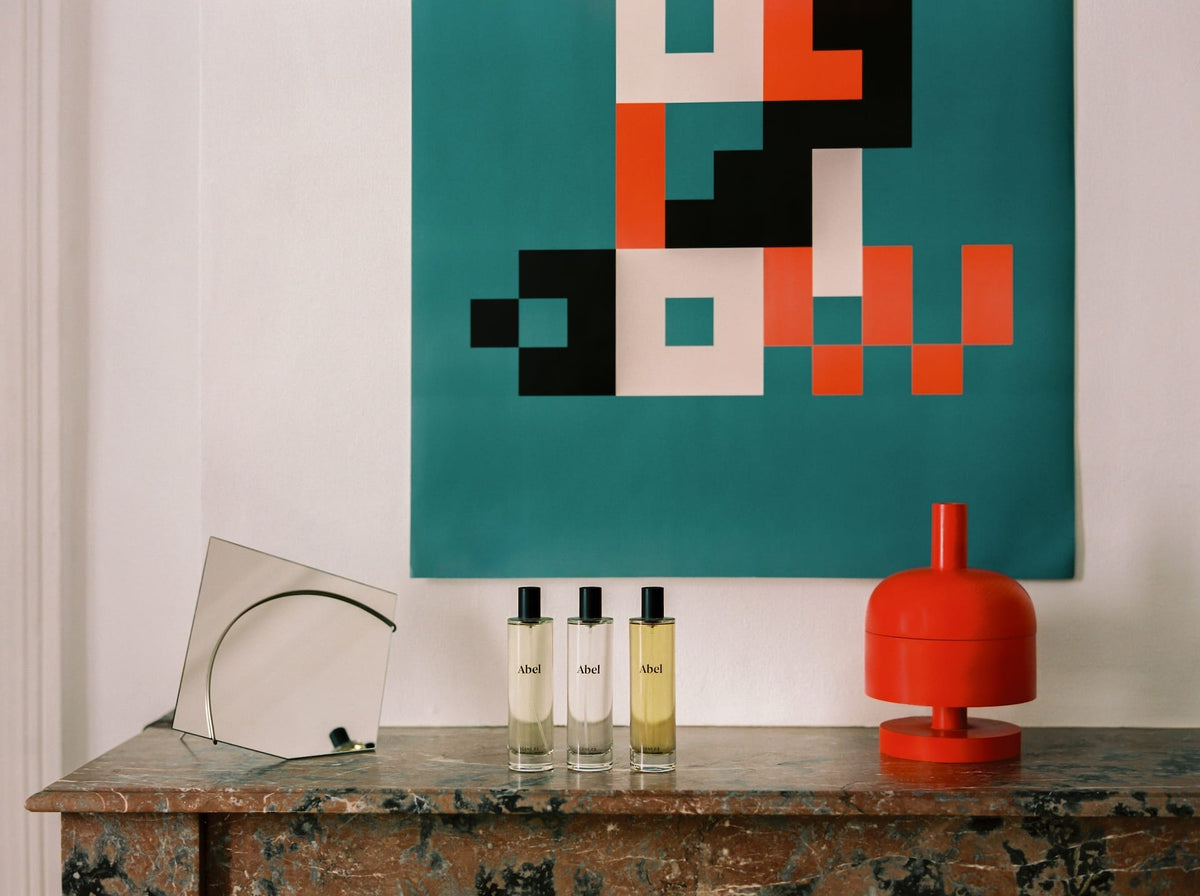 A minimalist interior setup featuring a transparent curved object, three Abel Room Spray – Scene 01 ⋅ green tea, yuzu, verbena bottles, and a red lamp on a marble surface, with a geometric abstract artwork in the background.