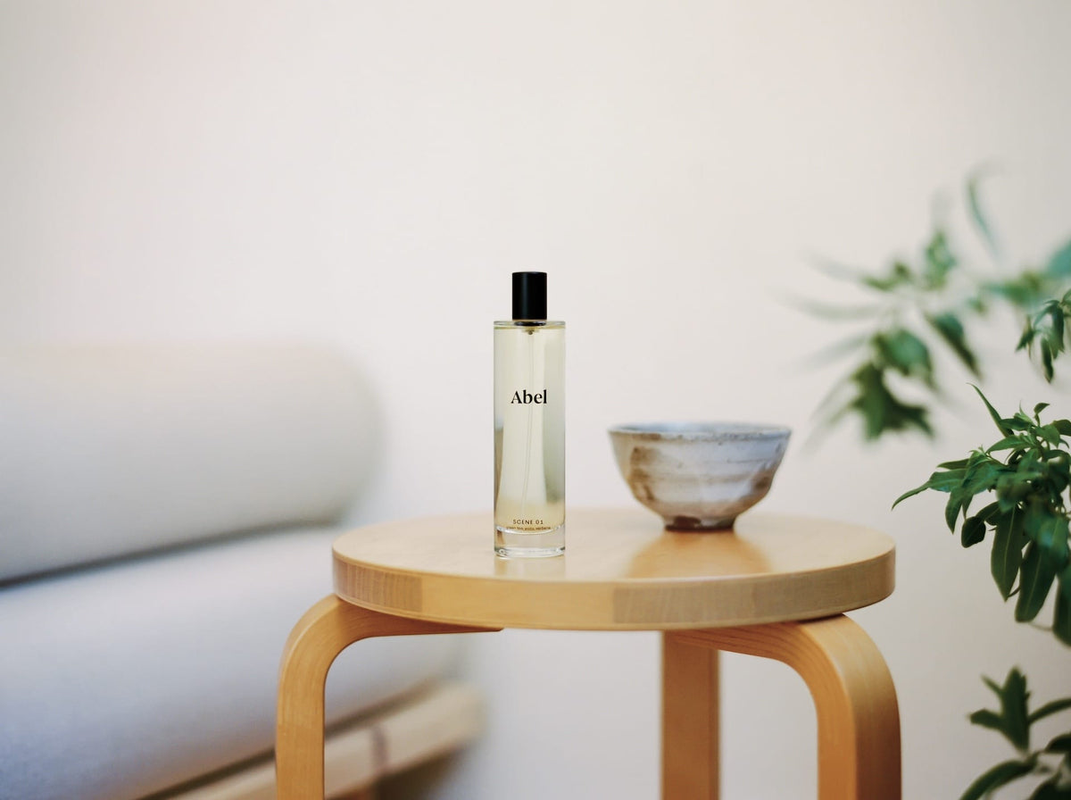 A bottle of Abel Room Spray – Scene 01 ⋅ green tea, yuzu, verbena on a wooden table beside a ceramic bowl, with a plant and part of a couch in the background.