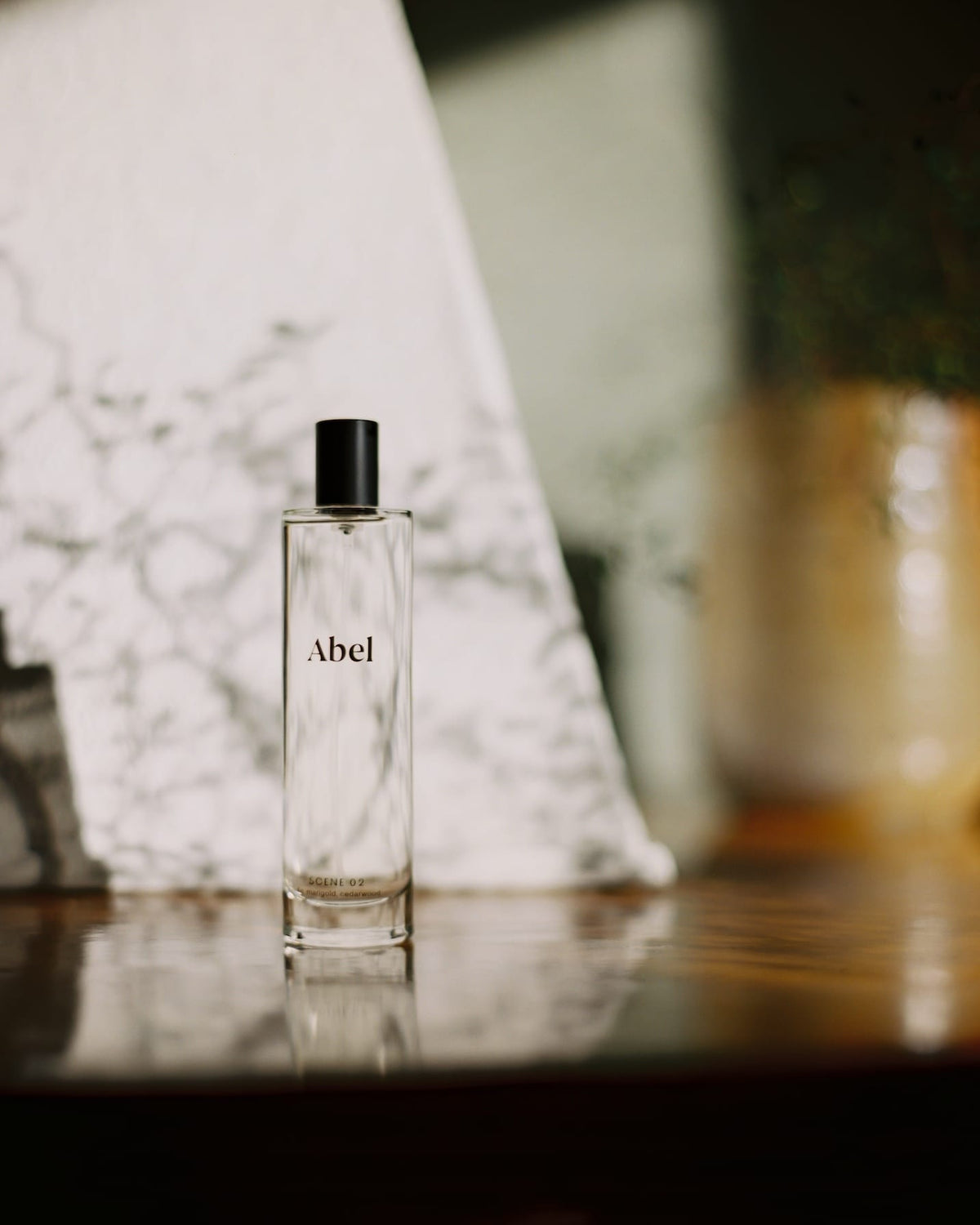 A bottle of Abel Room Spray - Scene 02 ⋅ fig, marigold, cedarwood on a reflective surface with a blurred background.