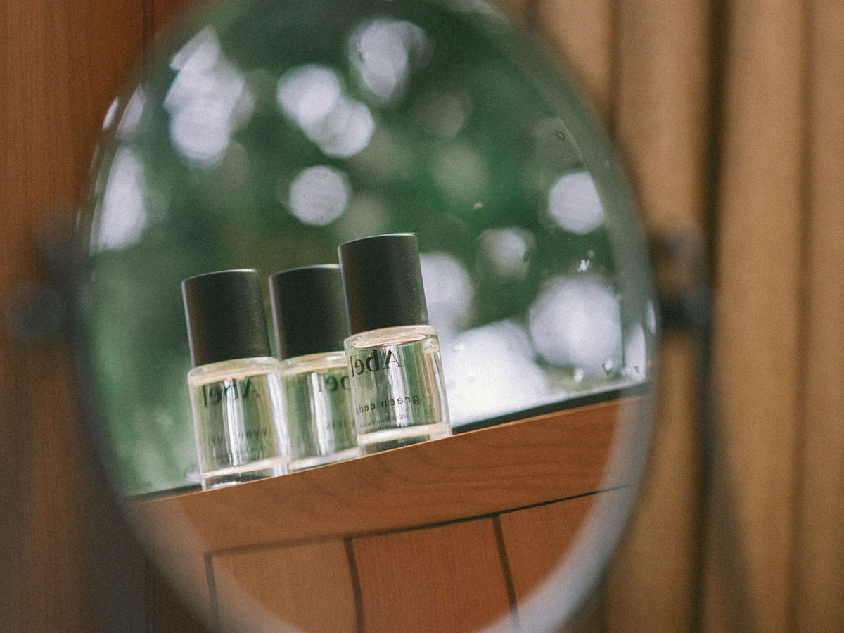 Three Layering Set – Cyan Nori ⋄ Green Cedar ⋄ Pink Iris eau de parfum bottles from Abel reflected in a round mirror with a blurred greenery background.