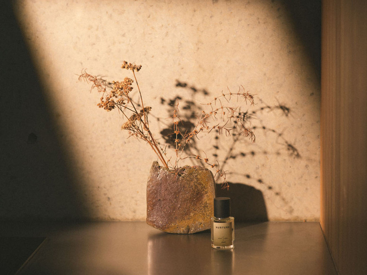 A bottle of NURTURE - grey label perfume by Abel sits on a table next to a vase of dried flowers emanating a delightful fragrance.