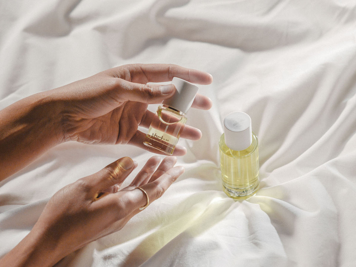 A person&#39;s hands applying perfume from a small bottle onto their wrist, with another bottle of Abel White Vetiver natural eau de parfum lying nearby on a wrinkled white fabric surface.