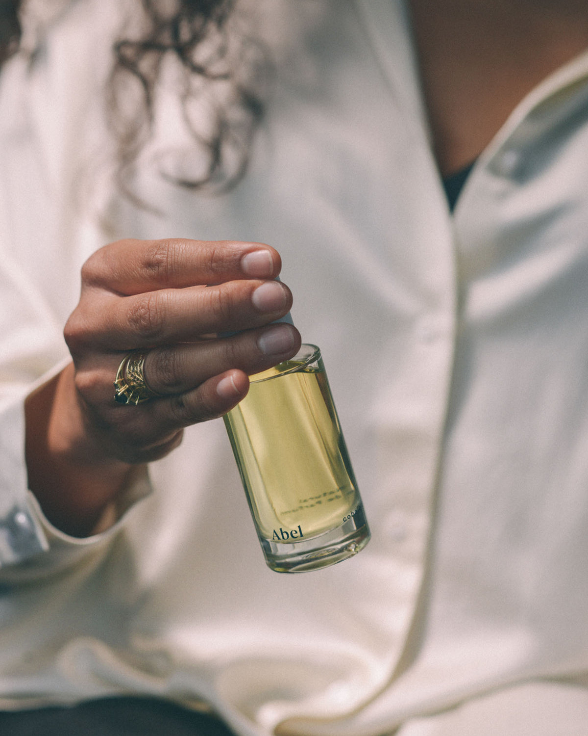 A sophisticated woman holding a small bottle of Cobalt Amber perfume scented with juniper berry and pink pepper, by Abel.