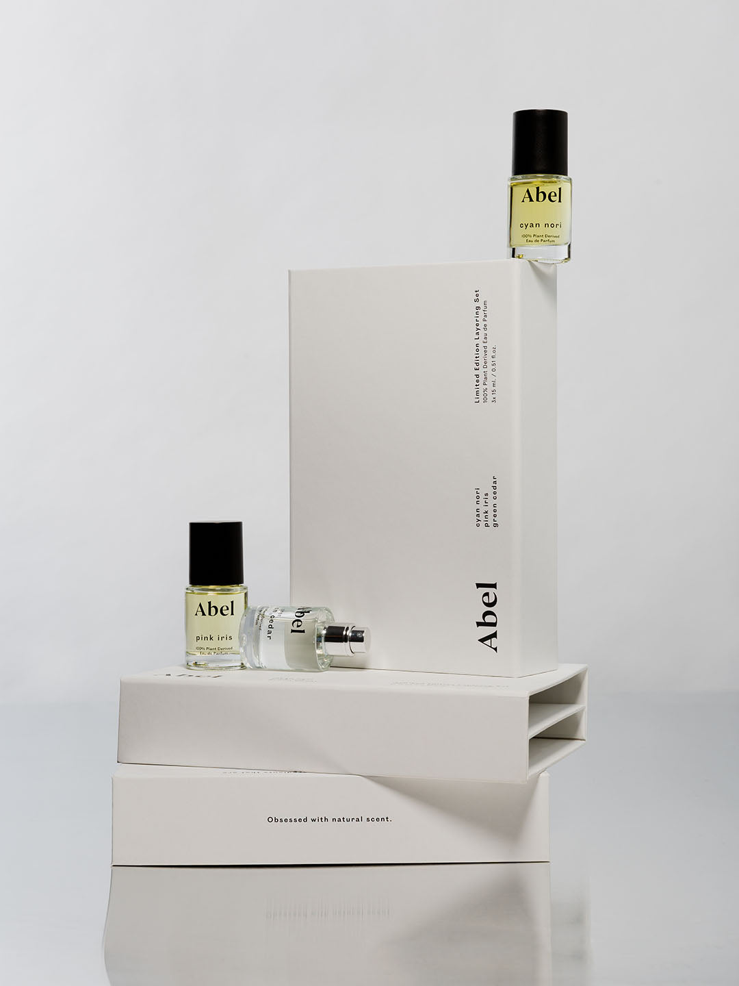 Two bottles of Abel Layering Set – Cyan Nori ⋄ Green Cedar ⋄ Pink Iris natural eau de parfum displayed on a minimalist white background, with a large and a small bottle in front of a rectangular pedestal, forming an elegant layering set