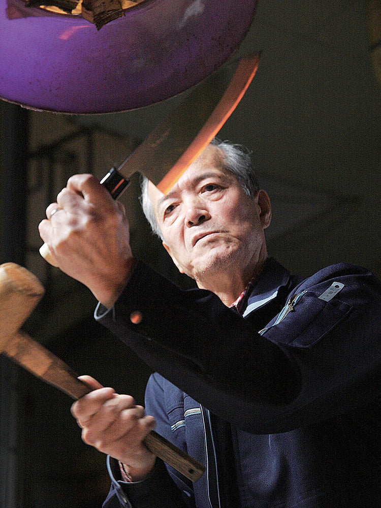 An elderly man skillfully sharpens a large knife using a hammer in a dimly lit workshop.