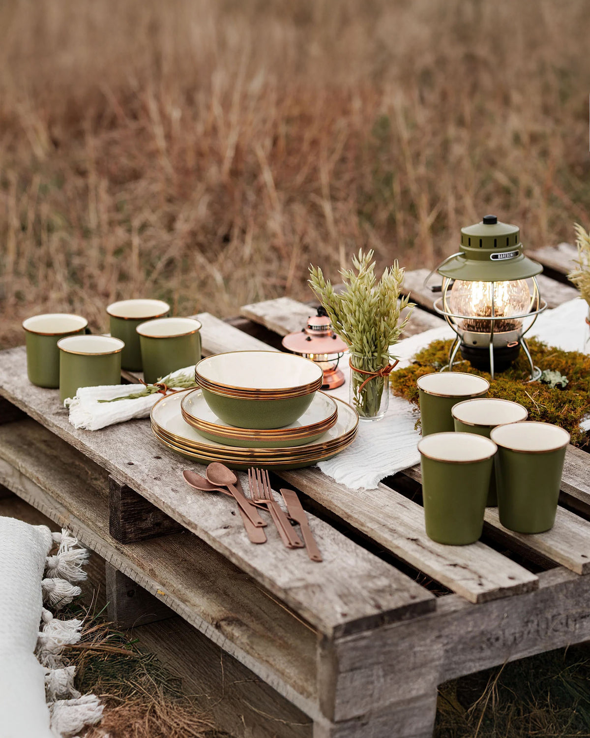 The outdoor table setup uses Barebones Enamel Two-Tone Tall Cups in Olive Drab, green enamelware plates, copper utensils, a lantern, and greenery. Its vintage-inspired design adds charm and seamlessly complements any garden party ambiance.