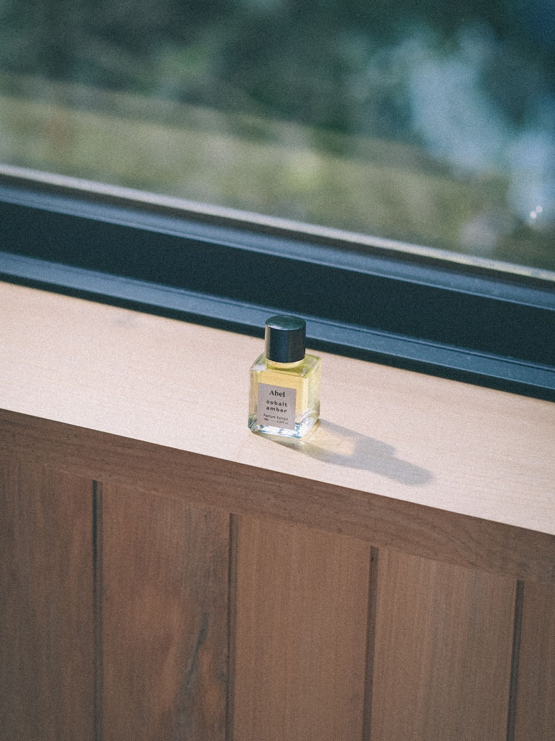 A travel-friendly small bottle of Cobalt Amber Parfum Extrait, enhanced with therapeutic-grade essential oils, elegantly placed on a window sill by Abel.