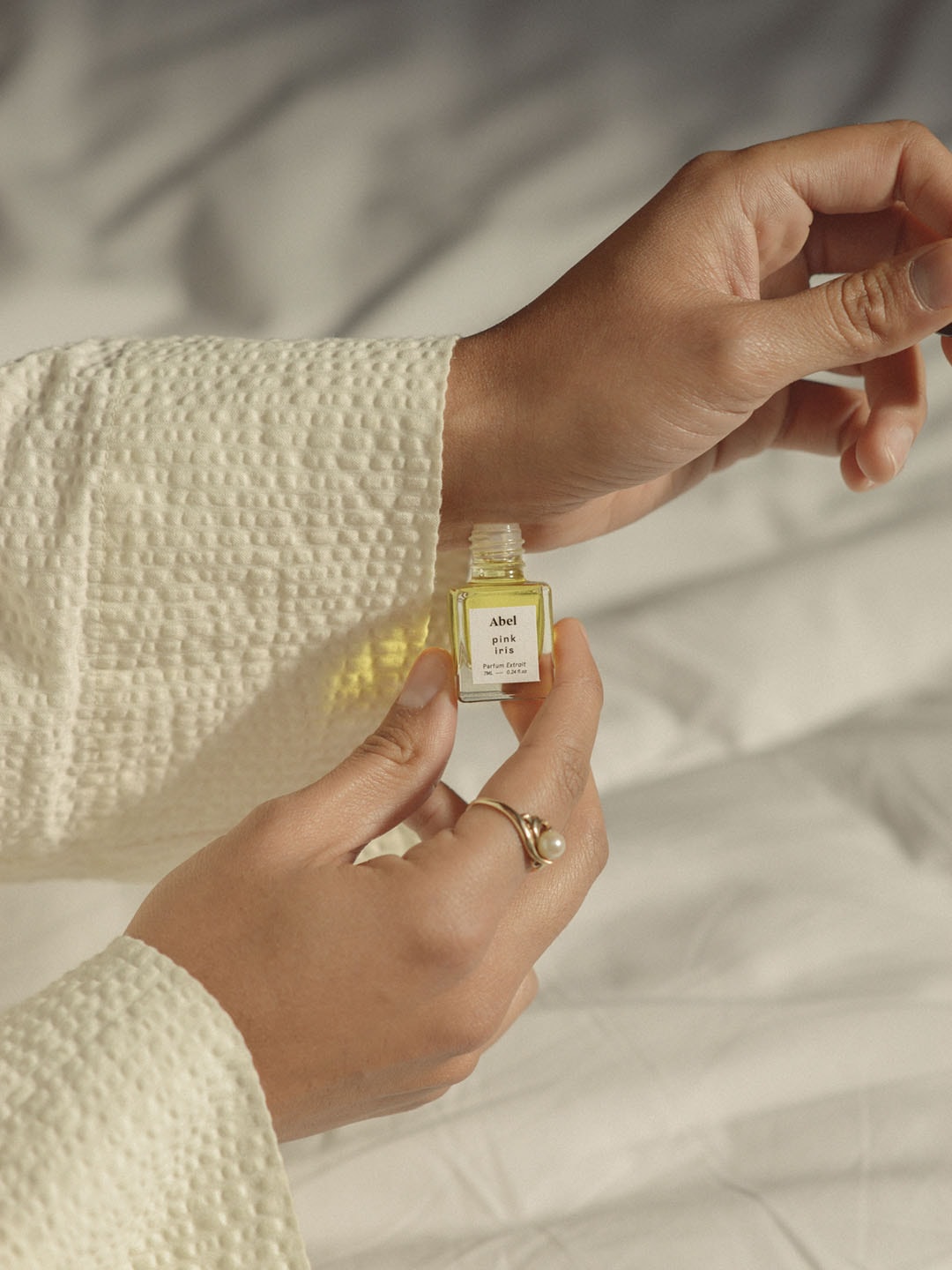 A woman holding a small bottle of Pink Iris Parfum Extrait by Abel on a white bed.