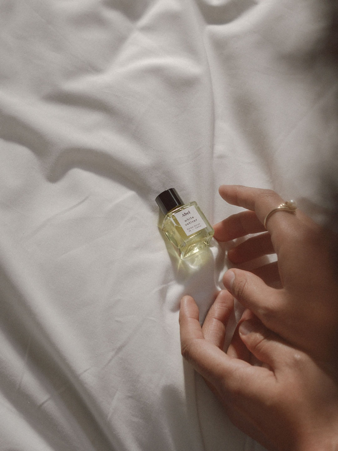 A hand holding a small bottle of White Vetiver Parfum Extrait perfume by Abel on a white bed.