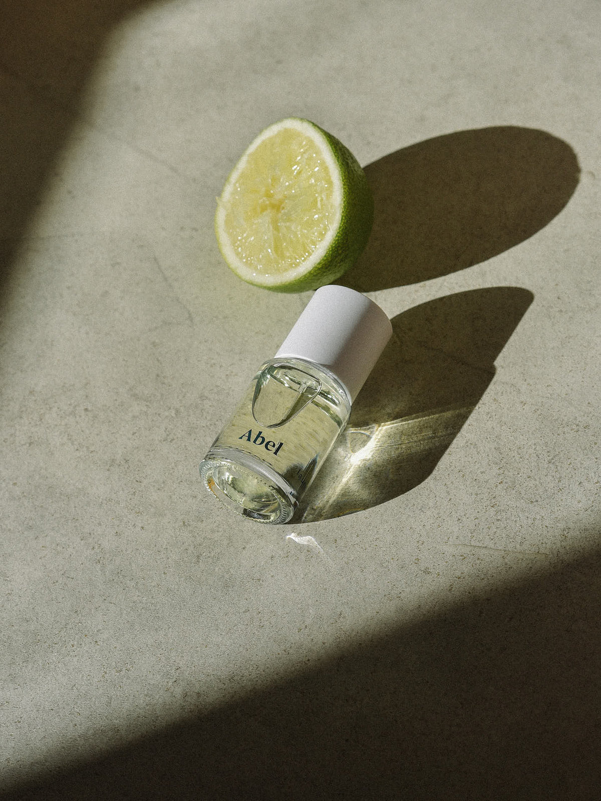 A perfume bottle labeled &quot;Abel&quot; rests on a concrete surface next to a halved lime, its shadows cast by sunlight. This 100% natural, citrus eau de parfum is ethically sourced, bringing together purity and elegance in every scent of Laundry Day by Abel – a verdant, sun-filled citrus.