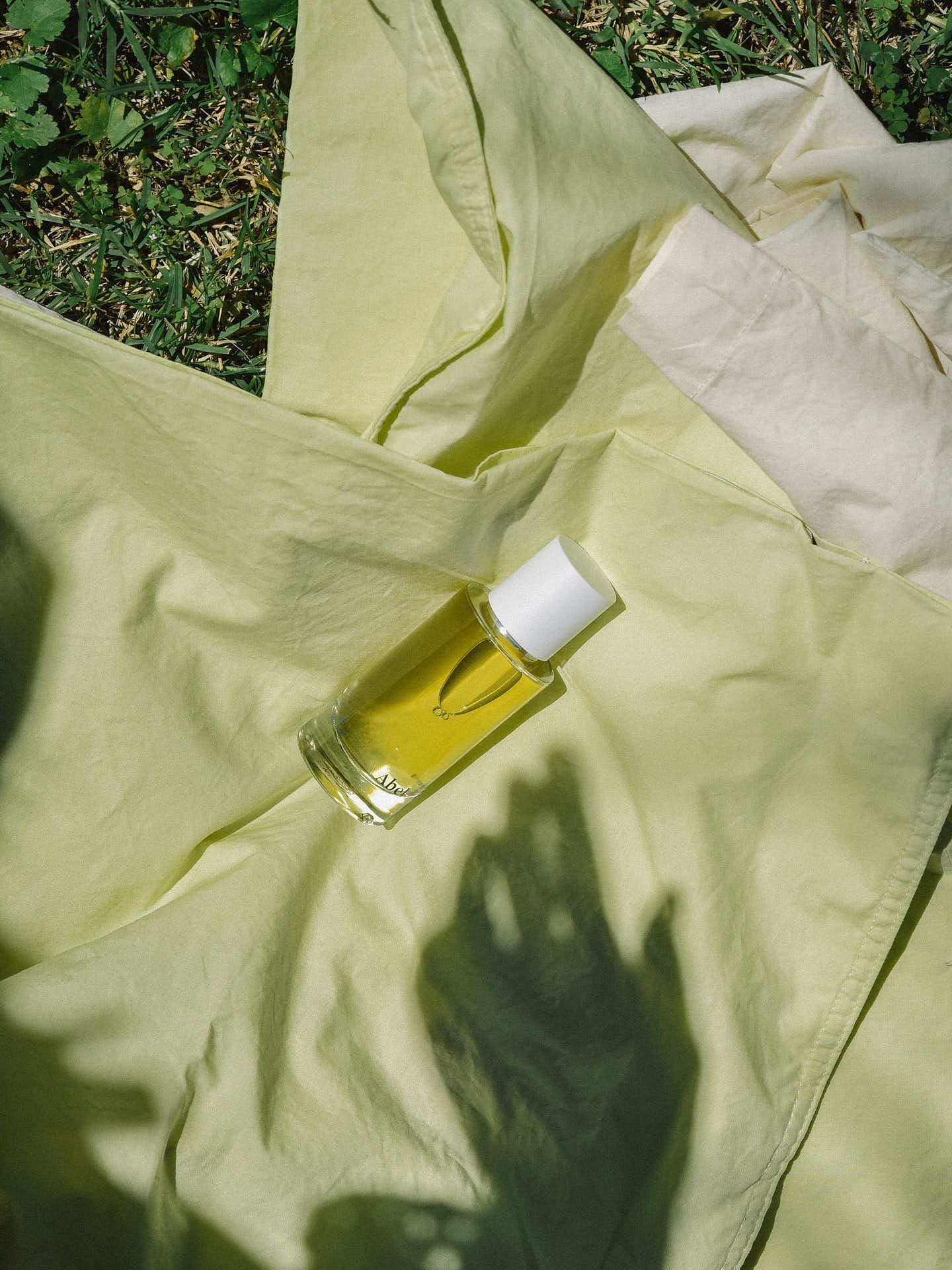 A clear glass bottle of Laundry Day – a verdant, sun-filled citrus by Abel with a white cap is placed on a light green cloth on grass, with shadows of hands visible on the cloth. The fragrance is 100% natural and ethically sourced.