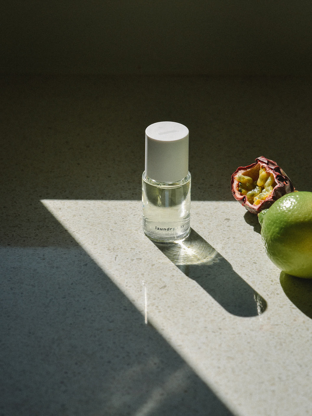 A small bottle of Abel’s Laundry Day – a verdant, sun-filled citrus rests on a stone surface beside a green lime and a halved passion fruit, lit by sunlight.