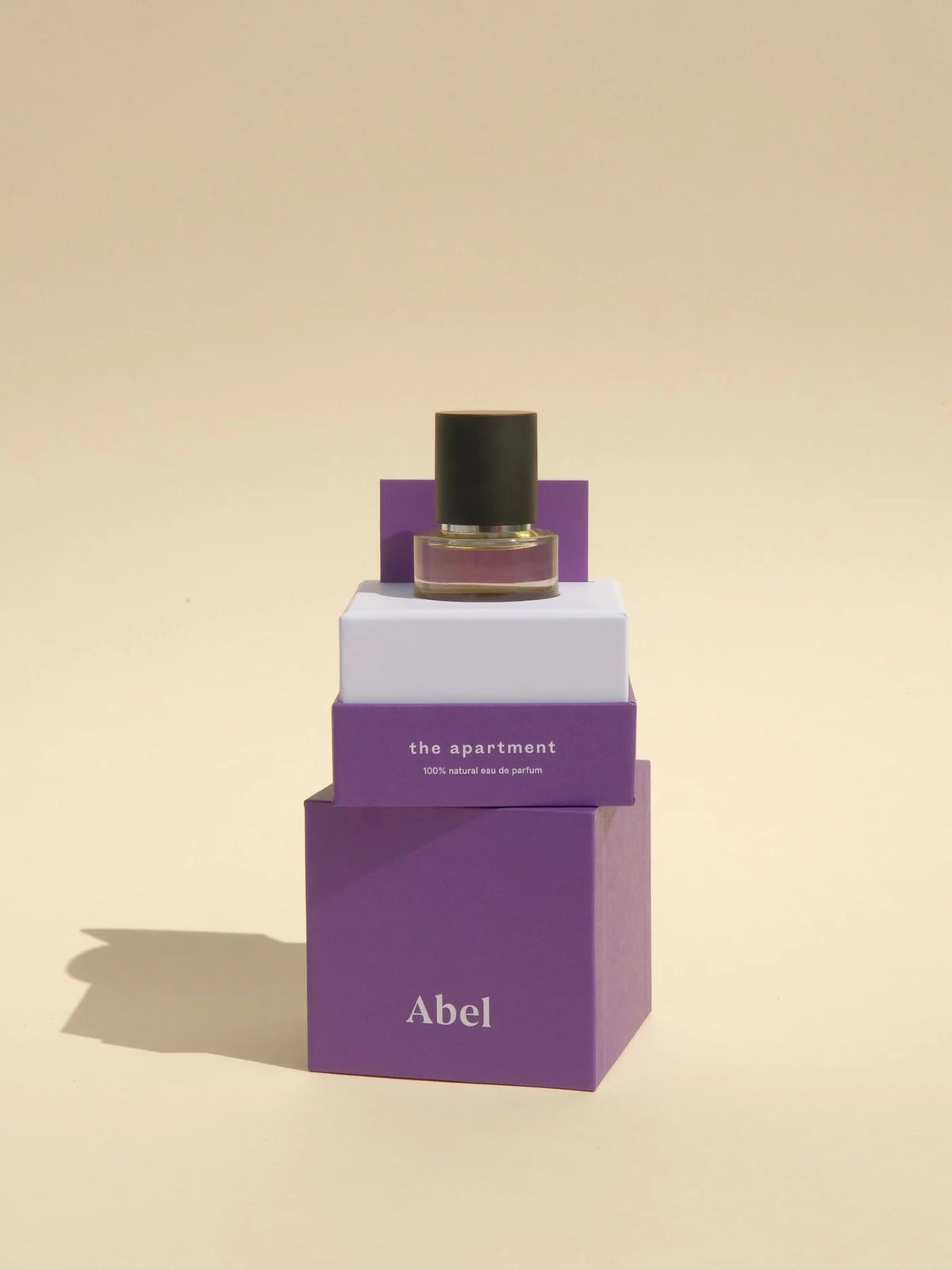 A luxurious bottle of fragrance from Abel&#39;s &quot;The Apartment&quot; collection rests elegantly on purple and white boxes, releasing the essence of a dark and sophisticated gourmand Eau de Parfum set against a beige background.